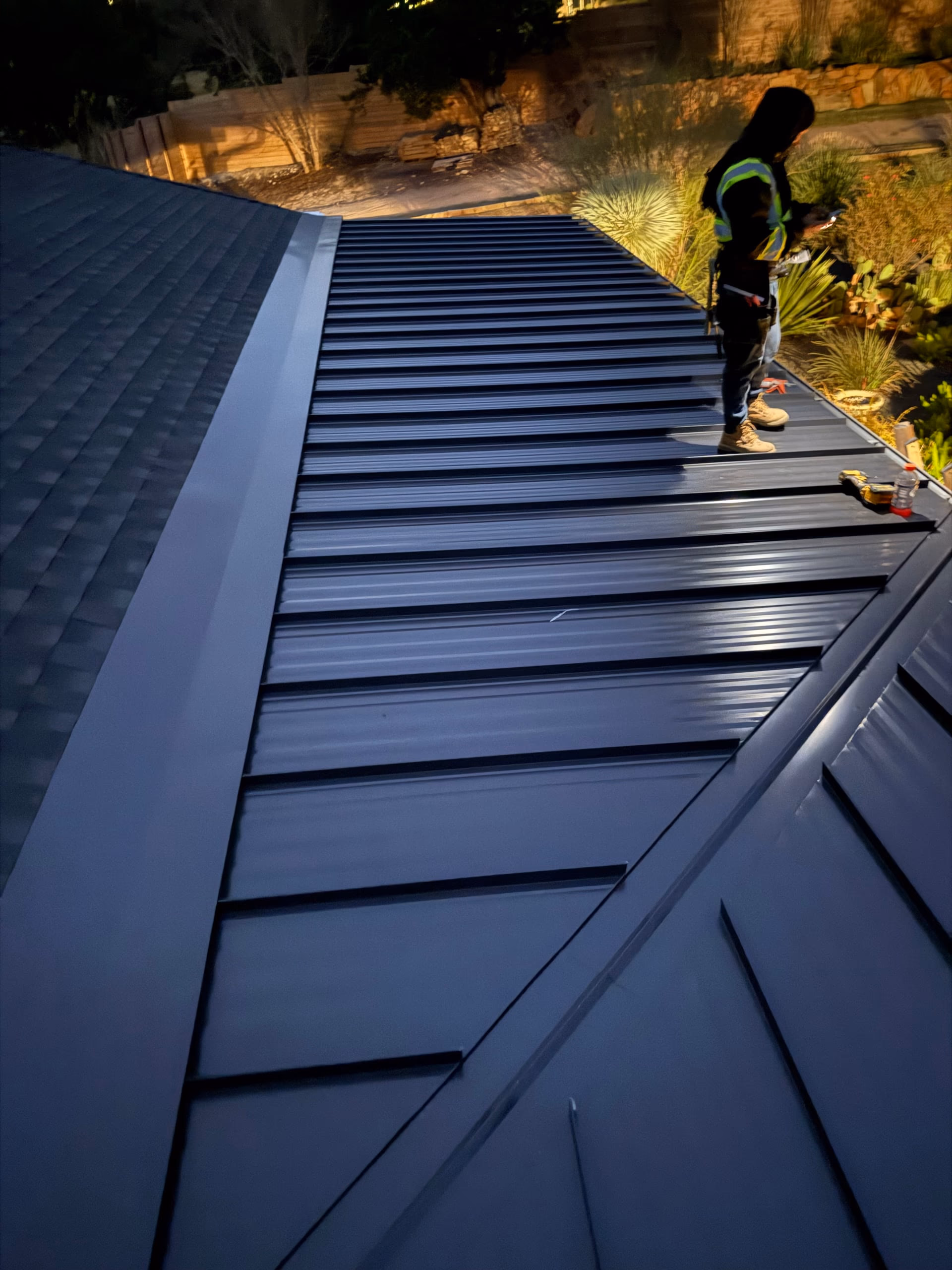 Newly installed dark bronze metal roof panels on a home at dusk with a roofer standing near the eave in Austin, TX