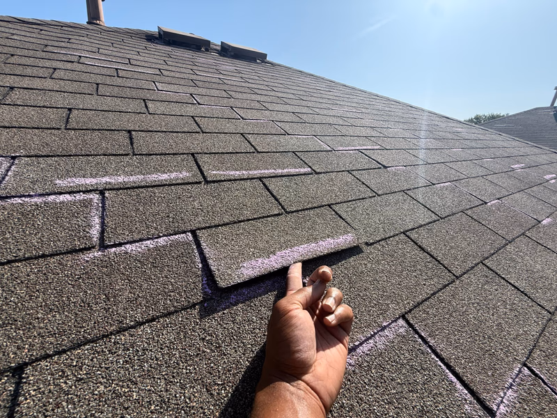 Hand lifting an unsealed wind damaged shingle tab with chalk inspection marks on an Austin, TX roof