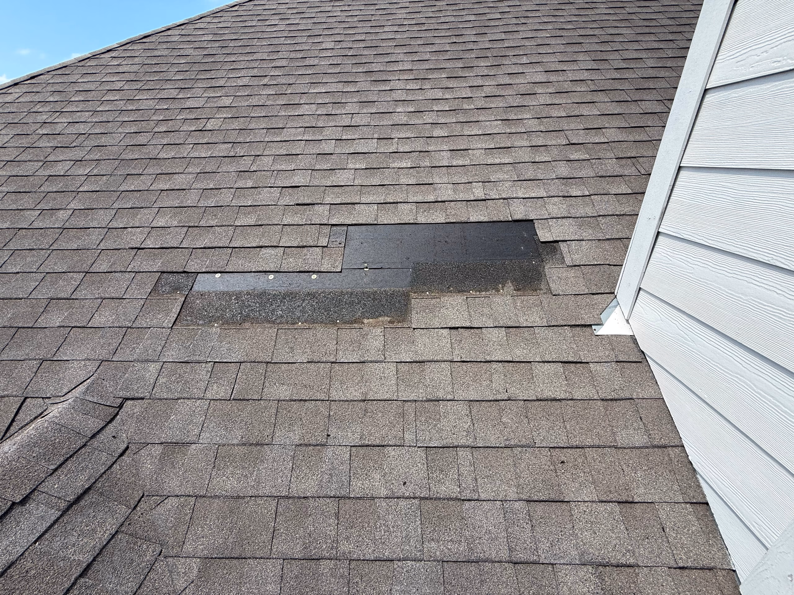 Section of missing shingles on a gray asphalt roof exposing black underlayment next to white siding on an Austin, TX home