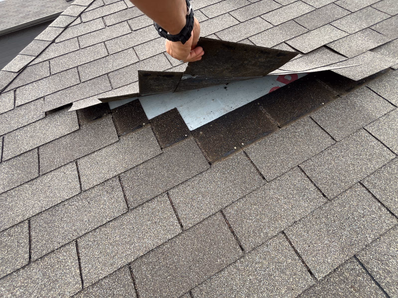 Roof inspector lifting a loose brown shingle by hand to check the underlayment beneath it during a free roof inspection in Austin, TX