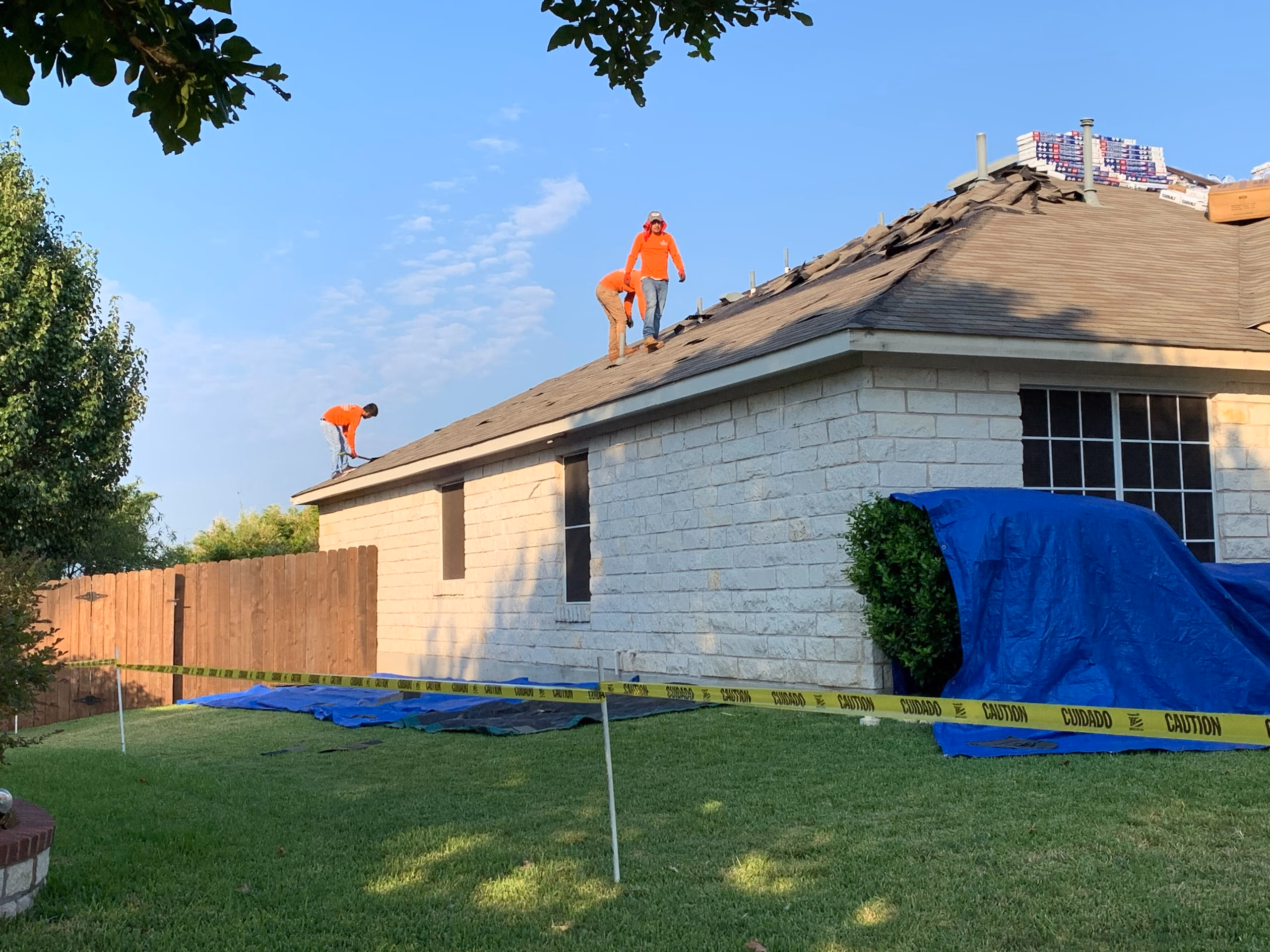Crew tearing old brown shingles off a stone home with tarps protecting the shrubs during a roof replacement in Austin, TX